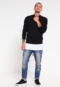 Svart långärmad tröja med rund halsringning, tillsammans med slitna blå jeans. Modell som bär svarta sneakers, står med neutral uttryck.