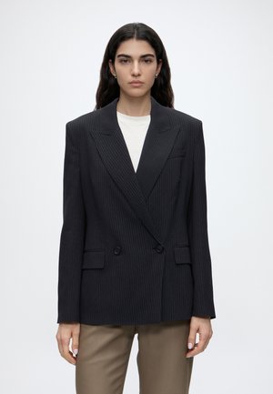 Donna dai lunghi capelli scuri indossa un blazer scuro a righe sottili sopra una camicia bianca e pantaloni color sabbia, in piedi contro uno sfondo chiaro e semplice.