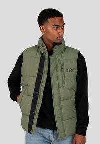Mann in grüner, gepolsterter Weste über einem schwarzen Sweatshirt und hellblauen Jeans, der zur Seite schaut, vor einem einfarbigen grauen Hintergrund.