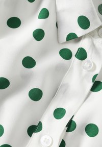 Blusa bianca con pois verdi, colletto con bottoni e una texture liscia. Presenta un bottone bianco vicino al colletto.