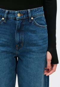 Τζιν παντελόνια με ψηλή μέση σε μπλε χρώμα, με αντίθετη ραφή, μπροστινές τσέπες και χρυσό κουμπί στο ύψος της μέσης. Ομαλή υφή υφάσματος.