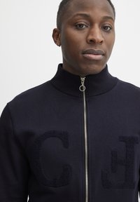 Marineblaue Zip-Jacke mit steilem Kragen, strukturiertem Buchstaben "C" und "J" auf der Vorderseite, aus glattem Stoff mit metallic Reißverschluss.