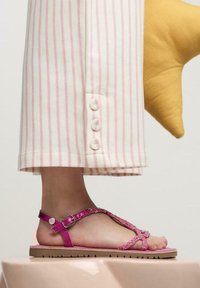 Sandalias rosas con tiras y un acabado texturizado, con detalles decorativos y una suela plana, combinadas con pantalones a rayas con detalles de botones.