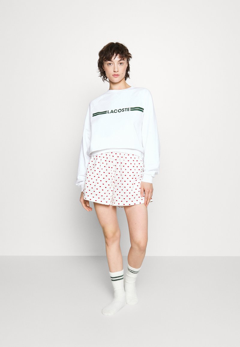 Sweat-shirt à manches longues blanc avec le texte "LACOSTE" en noir et vert, assorti de shorts blancs avec des motifs de cœurs rouges et des chaussettes rayées.