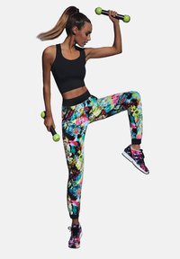 Czarny krótki top, kolorowe legginsy w kwiaty i wzorzyste trampki. Trzymając jasnozielone ciężarki. Dynamiczna poza na białym tle.