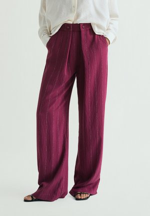 Persona que lleva pantalones magenta de talle alto y pierna ancha con sutiles rayas verticales, manos en los bolsillos, combinados con una blusa blanca y sandalias negras.