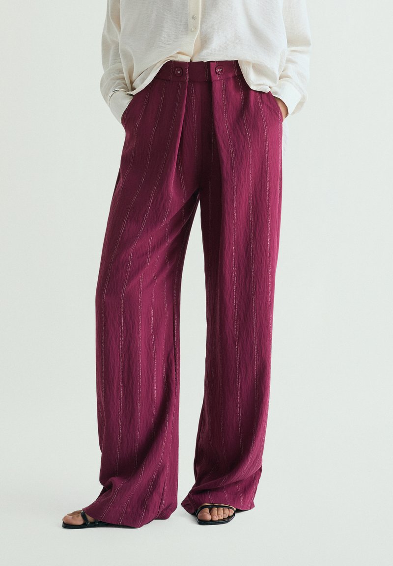 Persona che indossa pantaloni magenta a vita alta e gamba larga con sottili righe verticali, mani nelle tasche, abbinati a una camicetta bianca e sandali neri.