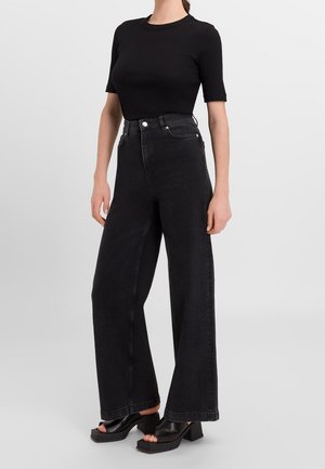 Osoba nosząca czarny dopasowany top z krótkim rękawem, czarne dżinsy z szerokimi nogawkami i wysokim stanem oraz czarne buty na słupku z odkrytymi palcami.