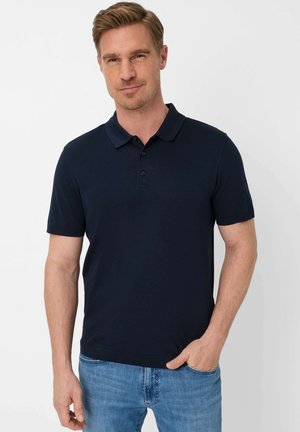 Man met lichtbruin haar, gekleed in een marineblauw poloshirt en lichtblauwe spijkerbroek, staand met één hand in de zak tegen een witte achtergrond.