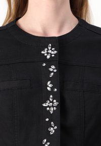 Indumento nero con un collo aderente, decorato con strass trasparenti disposti a grappolo a forma di fiori lungo la parte frontale.