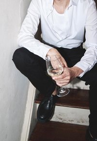 Zwarte leren schoenen, zwarte broek, wit overhemd met knopen, zittend op de trap, met een glas witte wijn in de hand. Neutrale muur en houten treden zichtbaar.