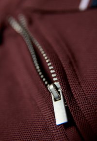 Bordowy materiał z srebrnym zamkiem; zbliżenie na suwak i zęby, ukazujące teksturowaną powierzchnię i detale szycia.