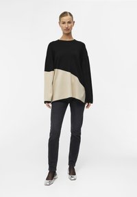 Maglione oversized nero e beige con un design a spacco diagonale, abbinato a jeans scuri e scarpe argentate, su uno sfondo neutro.