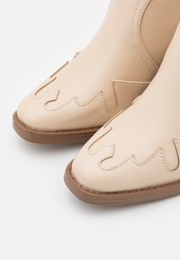 Botas de cuero beige hasta el tobillo con punta afilada, que cuentan con recortes en forma de ondas y un detalle de costura en contraste sobre una suela marrón.