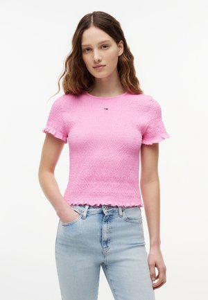 Młoda kobieta ubrana w dopasowany różowy top z krótkim rękawem o fakturowanej powierzchni oraz jasnoniebieskie, wysokowalne dżinsy, stojąca z jedną ręką w kieszeni.