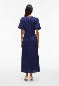 Žena v námořnickém midi šatech s vypouklými rukávy a bílými vysokými sandály, viděná zezadu na bílém pozadí.