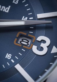 Cadran de montre analogique avec un fond bleu, des repères horaires blancs, des aiguilles argentées et une fenêtre de date bordée d'orange affichant le nombre 8.