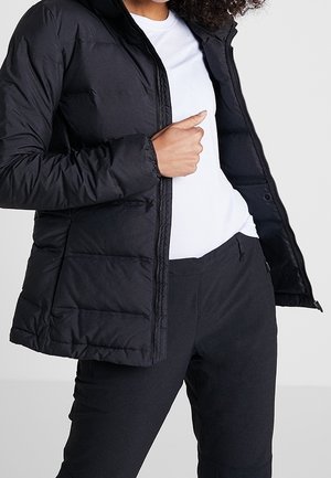 Personne portant une veste matelassée noire sur une chemise blanche et un pantalon noir, tenant la veste ouverte d'une main.