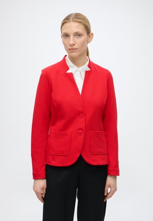 Vrouw met blond haar in een lage paardenstaart draagt een rode gebuttonede blazer over een wit overhemd en zwarte broek tegen een effen lichte achtergrond.