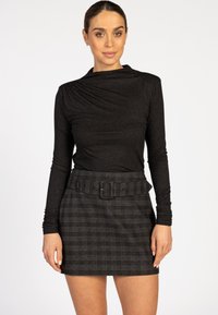 Vrouw in een zwart langoeve trui en een donkere geruite rok met een brede riem, staand voor een effen witte achtergrond.