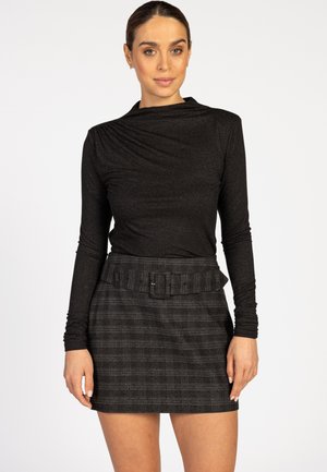 Woman wearing a black long-sleeve top and a dark plaid skirt with a wide belt, standing against a plain white background.