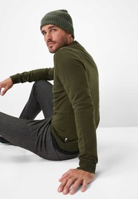Camicia verde a maniche lunghe e berretto a coste, abbinati a pantaloni grigi. La camicia presenta un collo rotondo e maniche aderenti. Tessuto liscio.