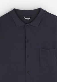 Polo shirt bleumarin cu guler, nasturi în față și țesătură texturată. Are un singur buzunar pe piept, pe partea stângă.