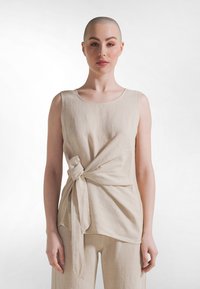 Beige mouwloze top van gestructureerde stof, met een knoopdetail aan de voorkant en een ontspannen pasvorm. Eenvoudige ronde halslijn en langere zoom aan de achterkant.