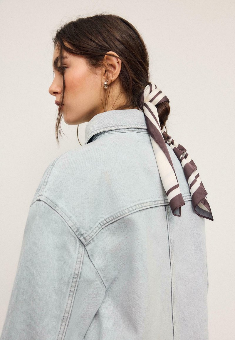 Jeune femme portant une veste en denim claire, cheveux foncés attachés en arrière avec un foulard rayé, tournée vers la gauche sur un fond uni.