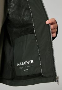 Sort zip-jakke med lynlås og mesh-fôr, indre label med teksten "ALLSAINTS" i hvid, og en glat ydre tekstur. Pyntet med reflekterende detaljer.