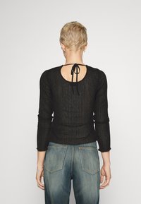 Schwarzes Langarmtop mit einem Schlüssel-Ausschnitt hinten und Bindedetail. Der Stoff ist transparent und hat ein strukturiertes Muster, kombiniert mit blauen Jeans.