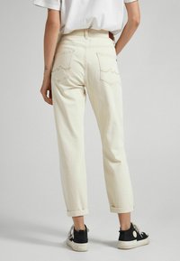 Henkilö, jolla on päällään vaaleanbeiget, käännetyt farkut, musta-valkoiset kävelykengät ja valkoinen T-paita, seisoo tasaisella vaalealla taustalla.