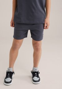 Shorts en coton gris avec une taille élastique et des poches latérales, associés à des chaussures de sport noires et grises arborant un logo sur la languette.