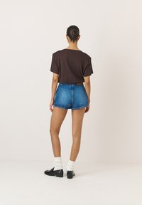 Tricou din bumbac maro asociat cu pantaloni scurți din denim albastri. Pantalonii scurți au margini desfăcute; modelul poartă pantofi negri și șosete albe cu textură ribbată.