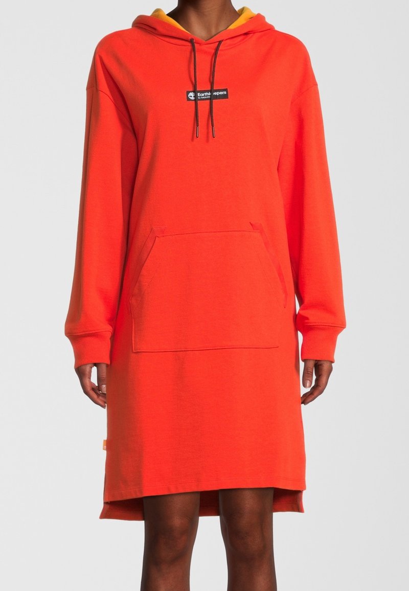 Personne portant une longue robe à capuche orange vif avec une poche avant et des cordons noirs, visible uniquement des épaules aux genoux.