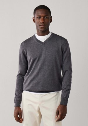 Joven con un suéter de cuello en V gris oscuro sobre una camisa blanca y pantalones beige claro, de pie frente a un fondo gris liso.