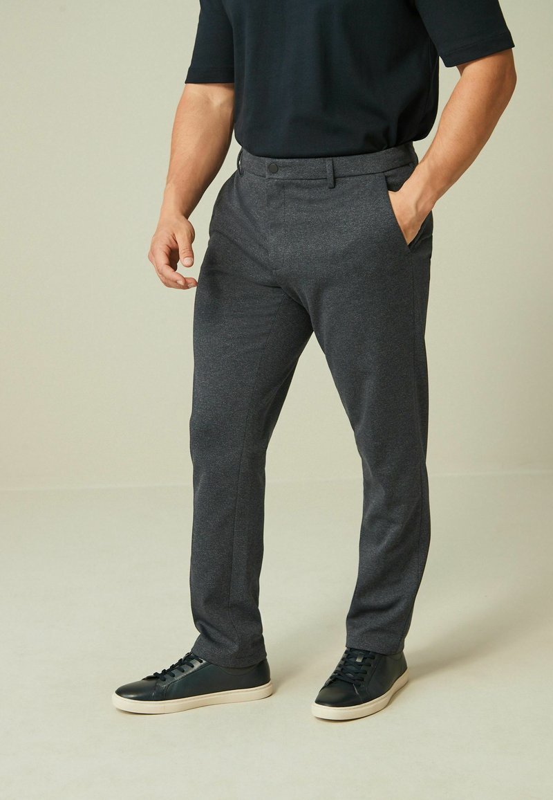 Graue maßgeschneiderte Hose aus glattem Stoff, mit flachem Frontdesign, seitlichen Taschen und schmaler Passform, kombiniert mit schwarzen Sneakern.