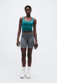 Αθλητική γυναίκα που φοράει πετρόλ αμάνικη μπλούζα, γκρι σορτς, λευκά αθλητικά παπούτσια, κρατώντας ένα παγούρι νερού, στέκεται μπροστά σε λευκό απλό φόντο.