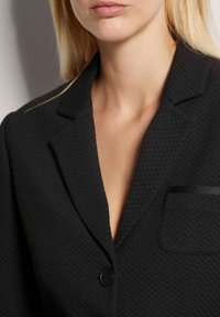 Schwarzer strukturierter Blazer mit Reverskragen, einem einzigen Knopfverschluss und einer kleinen Tasche mit einem schwarzen Akzentband.