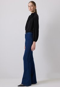 Top nero a maniche lunghe con fronte drappeggiato e dettagli floreali, abbinato a jeans svasati blu. La donna si trova di lato indossando un outfit aderente e scarpe a punta.
