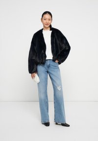 Giacca corta in pelliccia sintetica nera con maniche ampie, abbinata a un top bianco e a jeans a gamba larga strappati di un blu chiaro. Tiene una piccola borsa bianca.