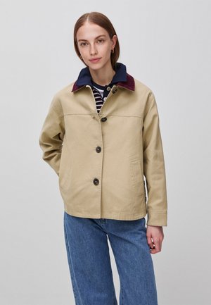 Vrouw draagt een beige jasje met knopen en een donkere kraag, over een gestreept shirt en blauwe spijkerbroek, staand tegen een effen lichte achtergrond.