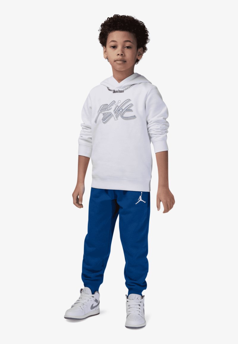 Sudadera blanca con logo plateado, pantalones de chándal azules con logo de Jordan y zapatillas blancas. La tela es suave; el corte es relajado y casual.