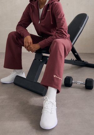 Person iført mørkerød træningsdragt og hvide sneakers, der sidder på en sort justerbar træningsbænk med en håndvægt på gulvet i nærheden.