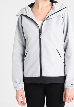Person trägt eine hellgraue Jacke mit Reißverschluss und schwarzem Besatz sowie verstellbaren Bündchen, kombiniert mit schwarzen Hosen vor einfachem Hintergrund.