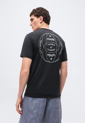 Mann trägt schwarzes T-Shirt mit weißem, rundem Grafik- und Textaufdruck "Roark", "The Beats of Discovery" und "Seek & Explore" auf der Rückseite, kombiniert mit grauen Shorts.