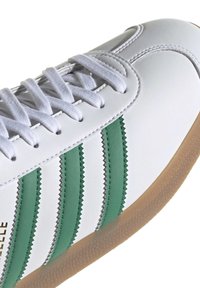 White leather sneaker with green serrated stripes, featuring a textured beige rubber sole and white flat laces.