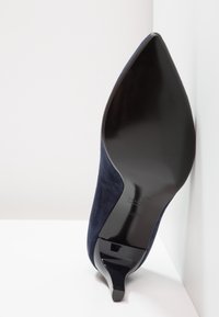 Escarpin en daim bleu marine avec un bout pointu et une semelle noire brillante, présentant un design latéral incurvé et une gravure de la marque sur la semelle.