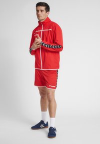 Rød atletisk jakke med hvide detaljer og sort chevronmønster på ærmerne, parret med røde shorts og blå sneakers. Hvide sokker synlige.