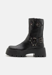 Botín de cuero negro hasta el tobillo con una suela gruesa, que presenta dos correas similares a cinturones con hebillas plateadas y un acento de anillo circular.